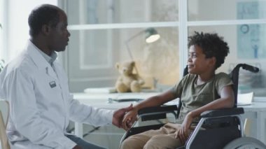 African American doctor speaking and holding hand on little boy in wheelchair during consultation in pediatric clinic