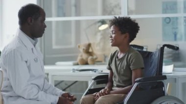African American doctor and boy in wheelchair talking and giving high fives in pediatric clinic
