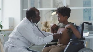 Psikiyatri kliniğinde sağlık kontrolü sırasında tekerlekli sandalyedeki çocuğu steteskopla muayene eden Afrikalı bir doktor.