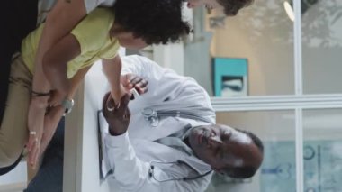 Vertical shot of African American doctor holding hand of little kid and talking to his mother during consultation in pediatric clinic