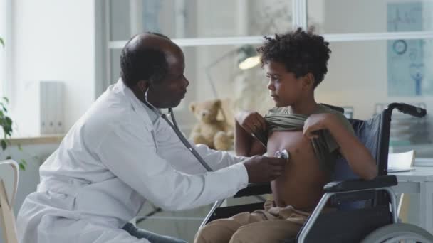 在儿科诊所进行体格检查时 非洲裔美国医生用听诊器检查坐在轮椅上的男孩 — 图库视频影像