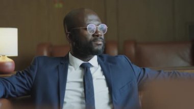 Portrait of African American businessman in formal suit and glasses sitting on leather sofa in hotel lobby and posing for camera