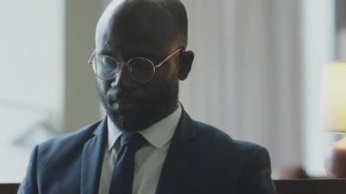 Tilt down shot of African American businessman in formal suit and glasses sitting on couch in hotel lobby and working on laptop