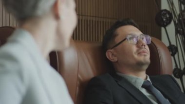Rack focus over the shoulder shot of businessman sitting in armchair and having talk with female colleague