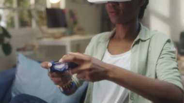VR kulaklıklı Afro-Amerikalı kadın evde sanal gerçeklikte oyun oynarken koltukta oturuyor ve oyun platformunu kullanıyor.