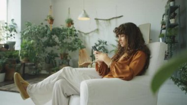 Oturma odasında yeşil çiçeklerle süslenmiş ve akıllı telefon kullanan genç ve güzel bir kadının görüntüsünü yakınlaştır.