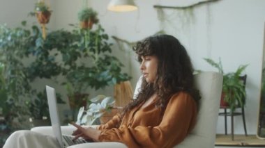 Genç bir kadının yeşil bitkilerle süslenmiş bir odada dizüstü bilgisayarla çalışırken orta ölçekli görüntüsü.