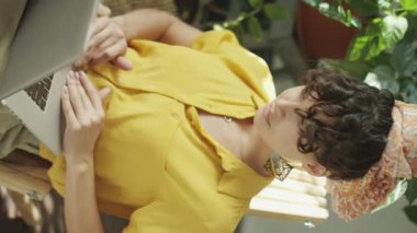 Genç bir kadının ev bitkileriyle aynı odada oturup dizüstü bilgisayarda yazışının dikey görüntüsü.