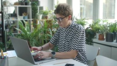 Genç iş kadınının dizüstü bilgisayar kullanırken ve iş günlerinde ev bitkileriyle süslenmiş rahat bir ofiste evrak işleriyle uğraşırken orta boy fotoğrafı.