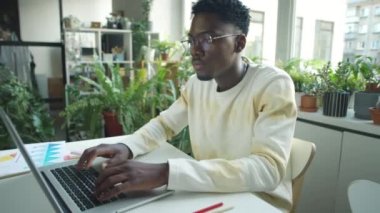 Genç siyahi iş adamının yeşil bitkilerle süslenmiş bir vaziyette masa başında dizüstü bilgisayarda daktilo kullanırken orta boy fotoğrafı.
