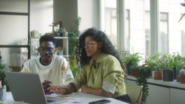 Latin iş kadınının, siyahi bir erkek meslektaşla birlikte rahat bir ofiste çalışırken dizüstü bilgisayarda proje tartışması.