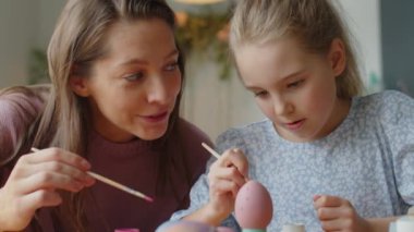 Neşeli anne ve küçük kız evde Paskalya dekoru yaparken boya fırçasıyla boyanmış yumurtalara noktalar çiziyorlar.