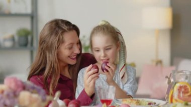 Neşeli anne ve küçük kız yemek masasında yumurta tokuşturup Paskalyayı evde kutluyorlar.