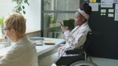 Tekerlekli sandalyedeki genç Afrikalı Amerikalı iş kadını ofiste çalışırken akıllı telefondan mesaj atıyor.