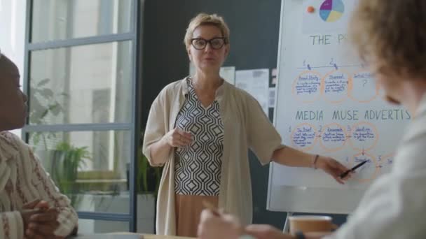 Bizneswoman Wskazując Schemat Flipchart Wyjaśniając Grupy Wielonarodowych Pracowników Podczas Szkolenia — Wideo stockowe