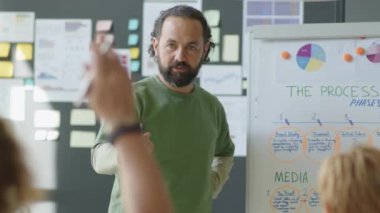 Erkek şirket eğitmeni, ofiste iş sunumu yaparken şemaya işaret ediyor ve çalışanlarla konuşuyor.
