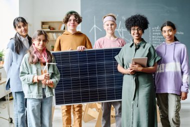 Öğretmenle birlikte güneş panelinin çalışmasını öğrenirken kameraya gülümseyen öğrencilerin portresi