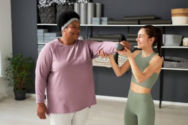 Sağlık kulübünde kıdemli tombul kadın çalıştıran beden eğitimi hocası, dambıllarla egzersiz yapıyorlar.