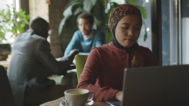 コーヒーショップのテーブルでラップトップに取り組むヒジャブのイスラム教徒女性の選択的な焦点ショット — ストック動画