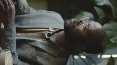 Afro-Amerikalı iş adamının bilgisayarda ve kahve masasında cappuccino içerken kameraya gülümseyişinin dikey portresi.