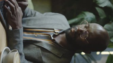 Resmi kıyafetli ve gözlüklü Afro-Amerikan işadamının kahve masasında dizüstü bilgisayarda uzaktan çekilmiş dikey görüntüsü.