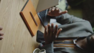 Kafe masasında dizüstü bilgisayar ve kahveyle oturan, el sıkışan ve iş arkadaşıyla konuşan Afro-Amerikan iş adamının dikey kesimi.
