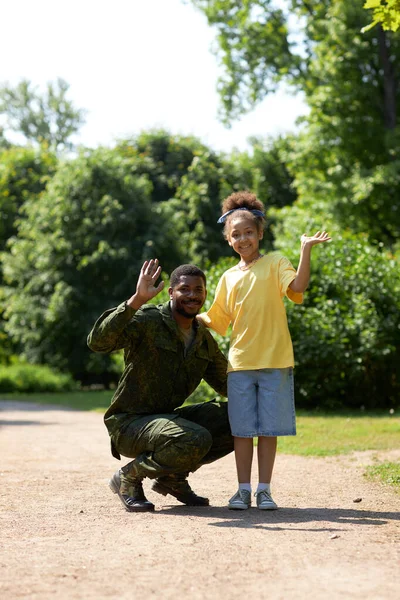 Portrait of African American dad in military uniform waving at camera together with his daughter
