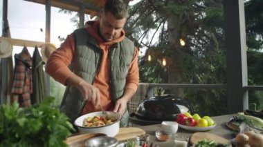 Erkek yemek blogcusu kasede sebzeleri karıştırıyor ve dışarıda barbekü yapmaya hazırlanırken kamerada konuşuyor.