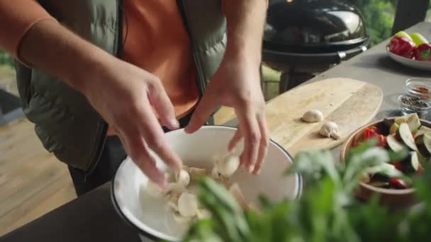 将男厨师的照片倾斜起来 将新鲜蔬菜和蘑菇放进碗里 在室外平台上做饭时对着镜头说话 — 图库视频影像