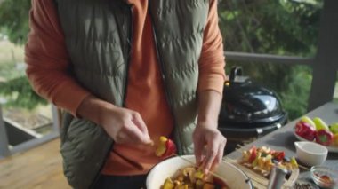 Erkek yemek blogcusu açık terasta bbq hazırlarken, şişte sebzeleri ipliyor ve kamerada konuşuyor.