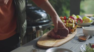 Açık terasta bbq pişirirken tahta tahtaya çiğ et koyan bir adamın kırpılmış görüntüsü.