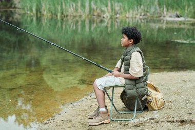 Çocuk elinde olta ve gölün yanında balık tutuyor.