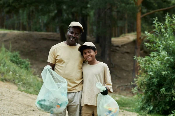 Portrait of African American dad and son smiling at camera while standing with garbage bags outdoors