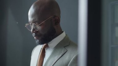 Resmi takım elbiseli ve gözlüklü Afro-Amerikan işadamının ofiste dikilip akıllı telefondan yazı yazarken çekilen görüntüleri.