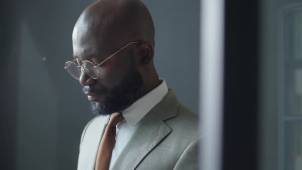 オフィスに立ってスマートフォンでタイピングする正式なスーツと眼鏡でアフリカ系アメリカ人のビジネスマンのチルトダウンショット — ストック動画