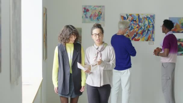 女性ギャラリーガイド 小冊子を持ち 現代美術展の周りに彼女を示しながら絵画について訪問者に伝える — ストック動画