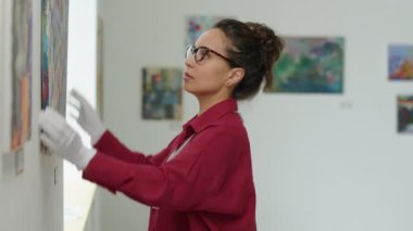 Beyaz eldivenli kadın galeri çalışanının sergi hazırlarken duvardaki resimleri kontrol ederken yan görüntüsü.
