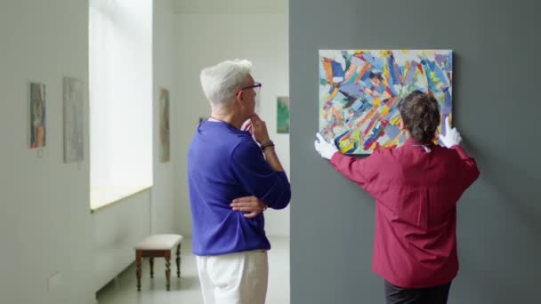 戴手套的女画廊工作人员在成年男性馆长的控制下将当代艺术挂在墙上 — 图库视频影像