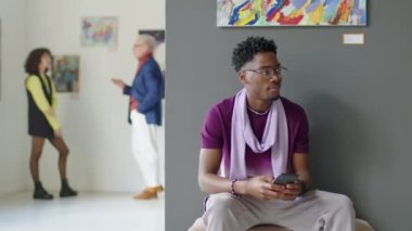 Genç Afro-Amerikan bir adamın akıllı telefon kullanıp sergi salonunda birini beklerken etrafa bakınırken görüntüsü.