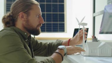 Erkek yenilenebilir enerji mühendisi ofiste ev ve rüzgar türbinlerinin modelleriyle çalışıyor.