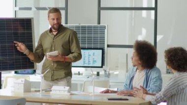 Yenilenebilir enerji mühendisi dijital tablet tutuyor ve ofis toplantısında ekibe sunum yaparken güneş panelini işaret ediyor