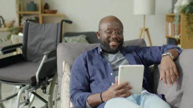 Tekerlekli sandalyenin yanındaki kanepede dinlenen özürlü Afrikalı Amerikalı bir adam ve evde boş zaman geçirirken tablet üzerinden görüntülü konuşma.