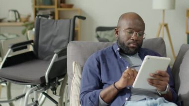 Engelli Afrikalı Amerikalı bir adamın tekerlekli sandalyenin yanında oturduğu ve evdeki oturma odasında dijital tablet kullandığı orta boy bir fotoğraf.