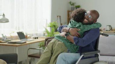 Evdeki oturma odasında tekerlekli sandalyede neşeli Afrikalı Amerikalı babaya sarılan küçük çocuk.