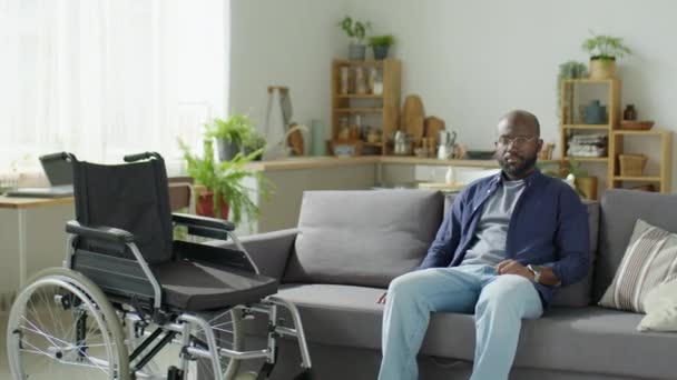 アフリカ系アメリカ人男性のアークショット 車椅子の横にソファーに座り 自宅で一日を過ごしながらカメラをポーズ — ストック動画
