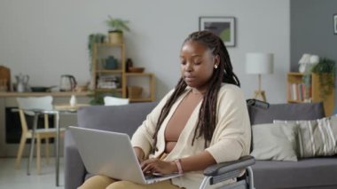 Özürlü genç Afrikalı Amerikalı kadın portresi tekerlekli sandalyede oturuyor, dizüstü bilgisayarını tutuyor ve oturma odasında kameraya poz veriyor.