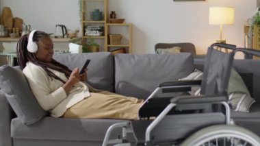 Engelli genç Afrikalı Amerikalı kadın tekerlekli sandalyenin arkasında dinleniyor ve evde vakit geçirirken akıllı telefondan kulaklıkla müzik dinliyor.