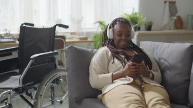 Engelli genç bir kadın tekerlekli sandalyenin yanındaki kanepede dinleniyor ve tüm gününü evde geçirirken akıllı telefonuyla müzik dinliyor.