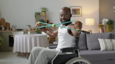 Orta boy, engelli Afrikalı Amerikalı bir adamın evde oturma odasında direniş bandıyla oturup antrenman yaptığı bir fotoğraf.
