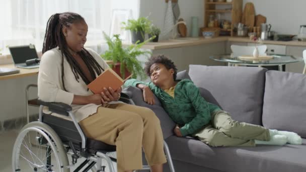 Afroamerikansk Mamma Rullstol Läser Bok Till Lille Son Kylning Soffan — Stockvideo